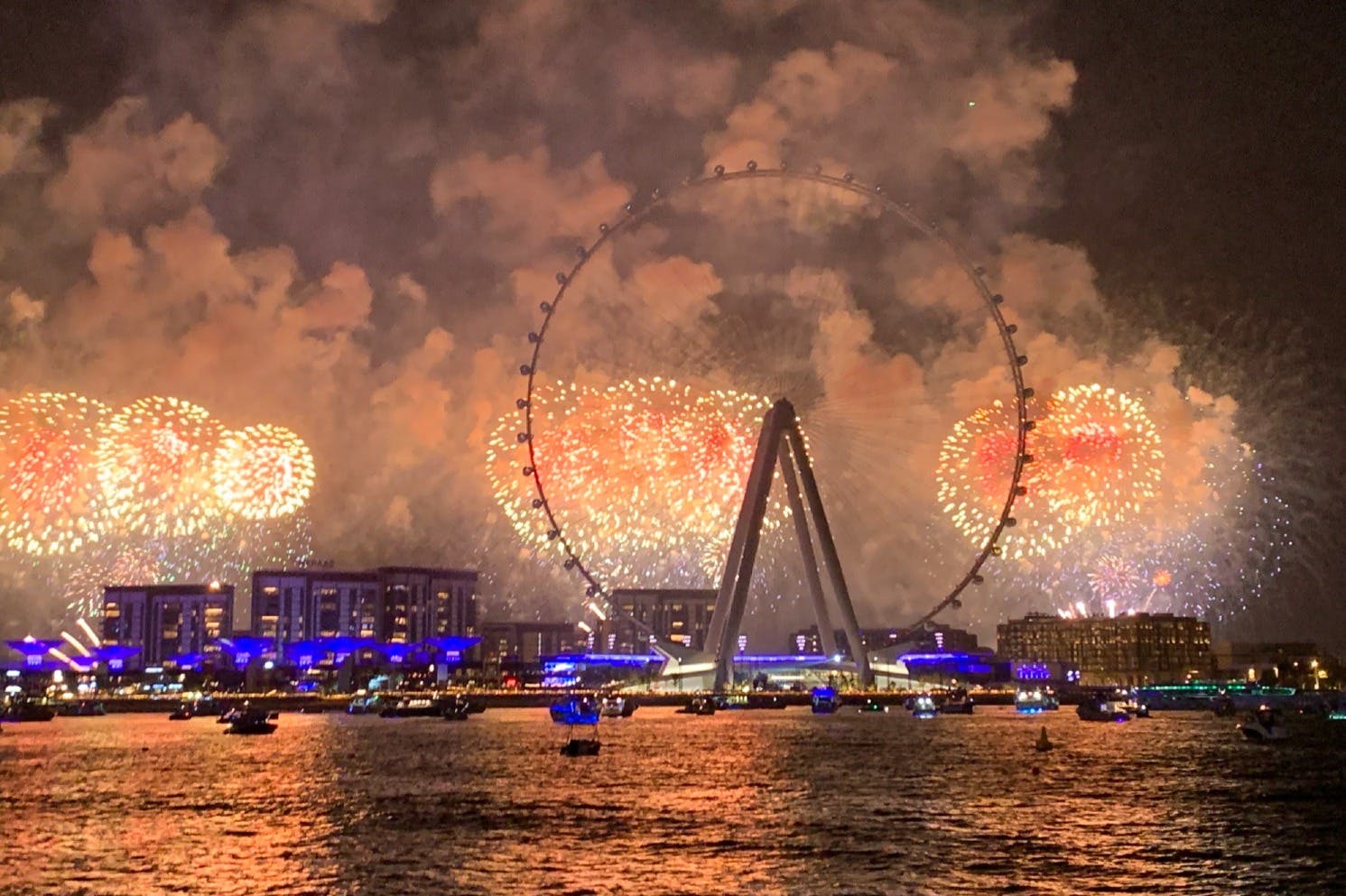 Riesenrad und Gebäude, beleuchtet von einem bunten Feuerwerk über dem nächtlichen Wasser, mit Booten im Vordergrund.