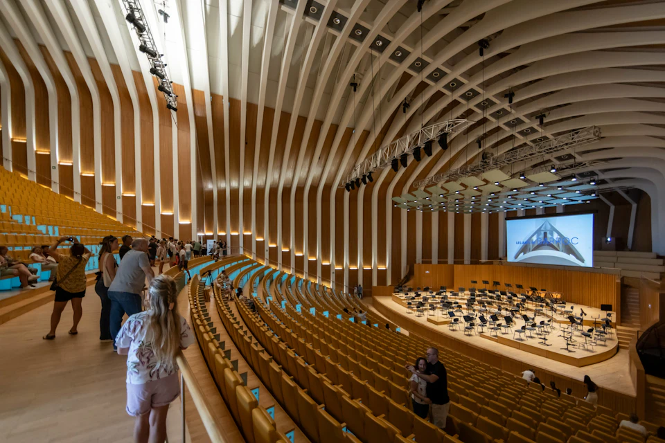 A modern concert hall with curved wooden walls, rows of empty yellow seats, and a stage set up for an orchestra performance.