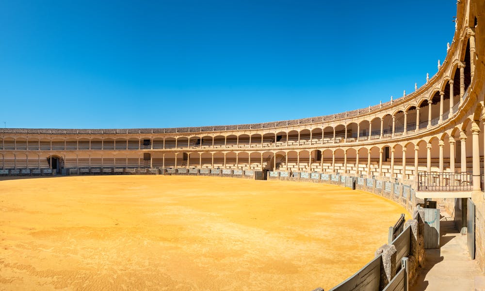 A large, empty bullring with sandy ground, surrounded by a two-tiered colonnade and arches under a clear blue sky.