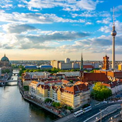 En stadsbild med en flod, historiska byggnader med röda tak, en katedral, ett TV-torn och en himmel fylld med spridda moln.