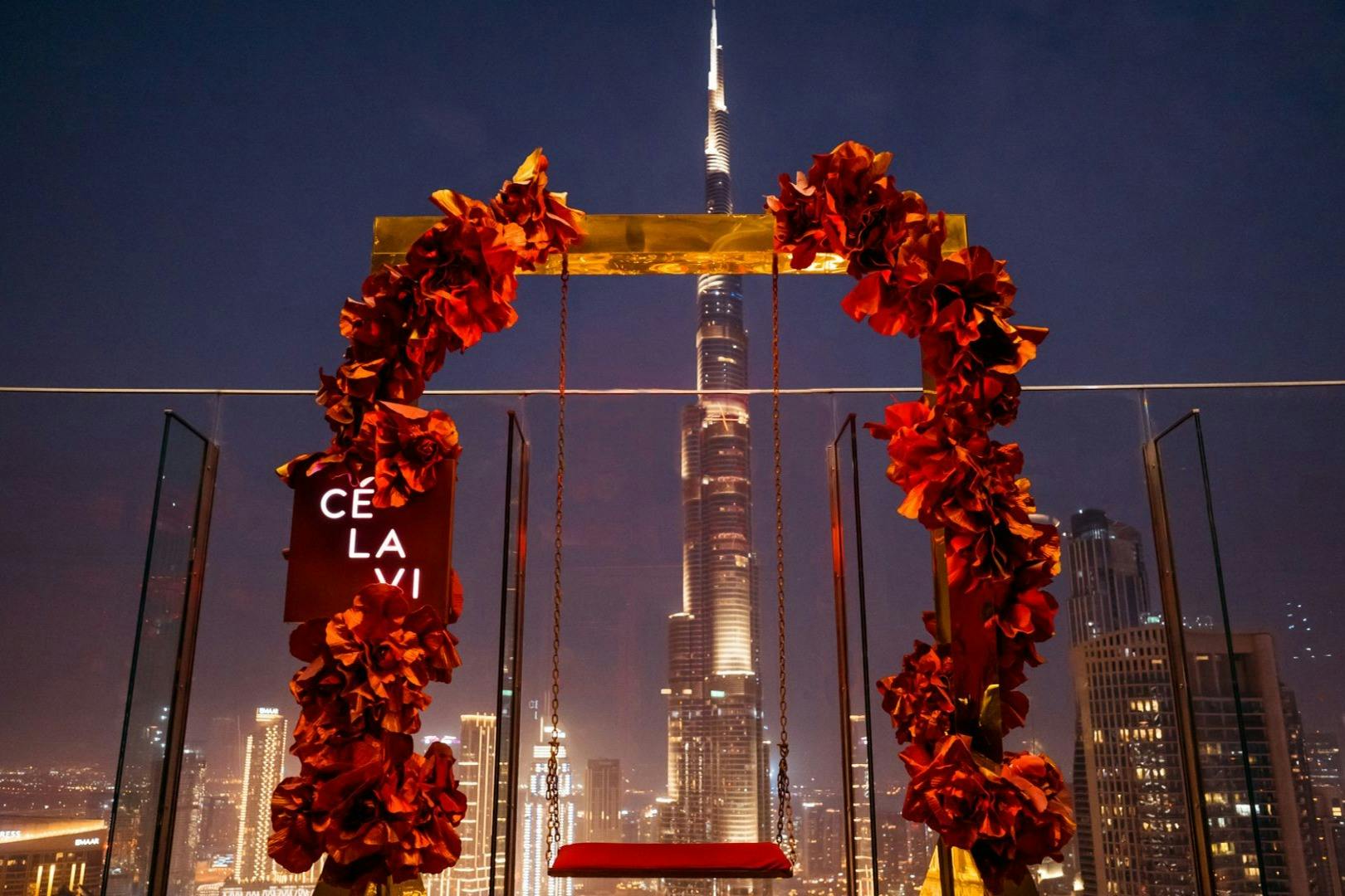 A swing decorated with red flowers in front of the Burj Khalifa at night. A sign on the left reads "CE LA VI."