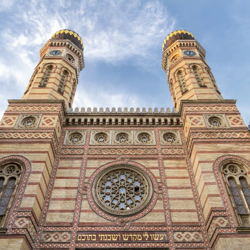 Utsmyckad fasad av en byggnad med två höga torn, invecklat tegelverk och hebreisk inskription, under en ljus, delvis molnig himmel.