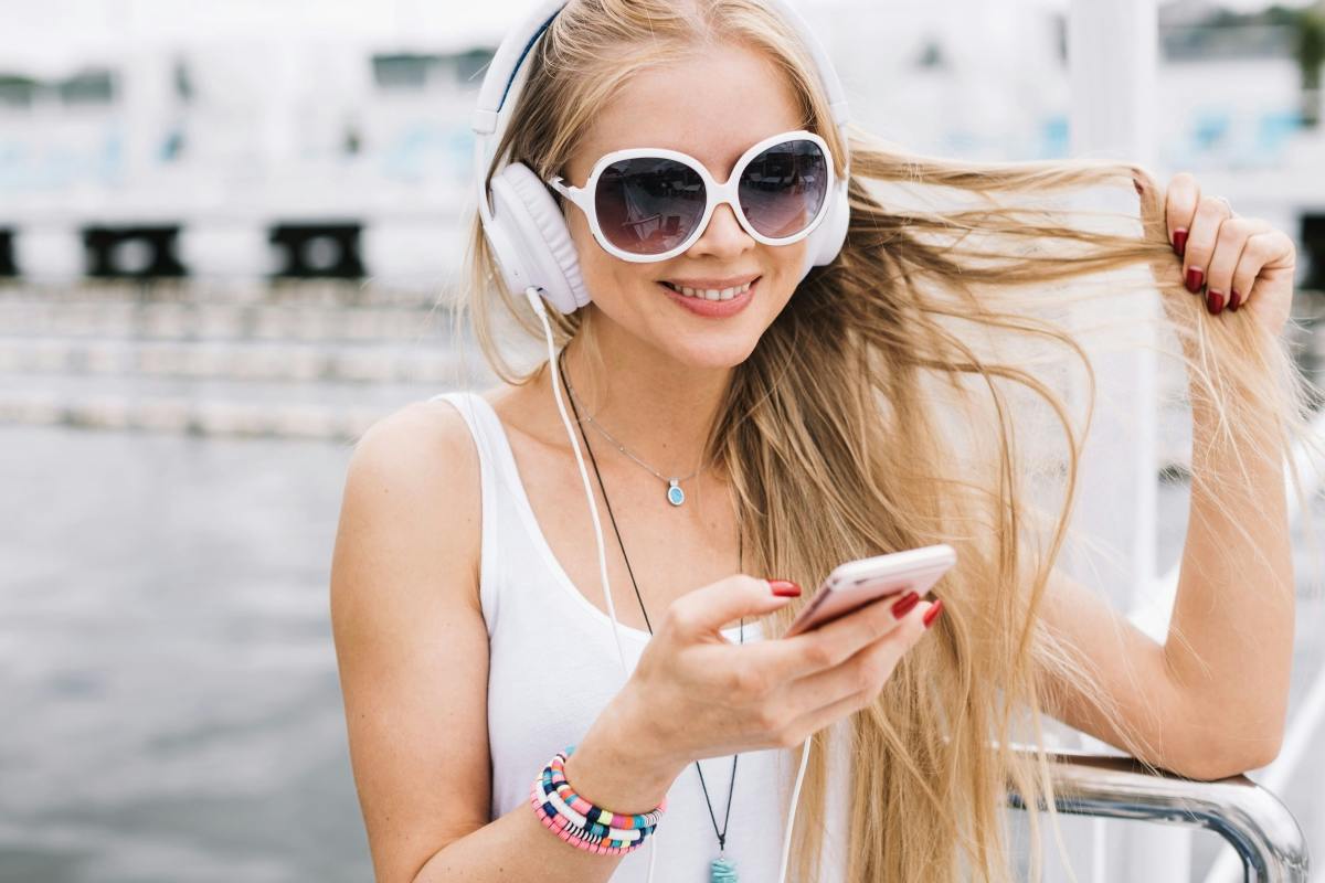 A woman wearing headphones and sunglasses holds a smartphone, smiling outdoors, with long hair flowing in the wind.