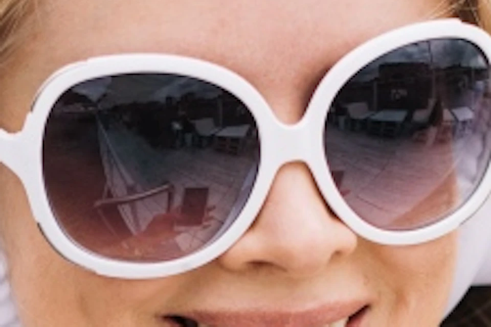 A woman wearing headphones and sunglasses holds a smartphone, smiling outdoors, with long hair flowing in the wind.