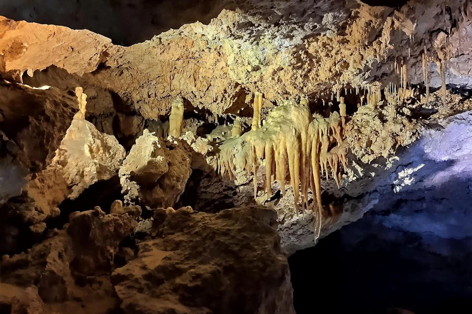 A cave interior with stalactites and stalagmites, illuminated by artificial lighting, showcasing textured rock formations.