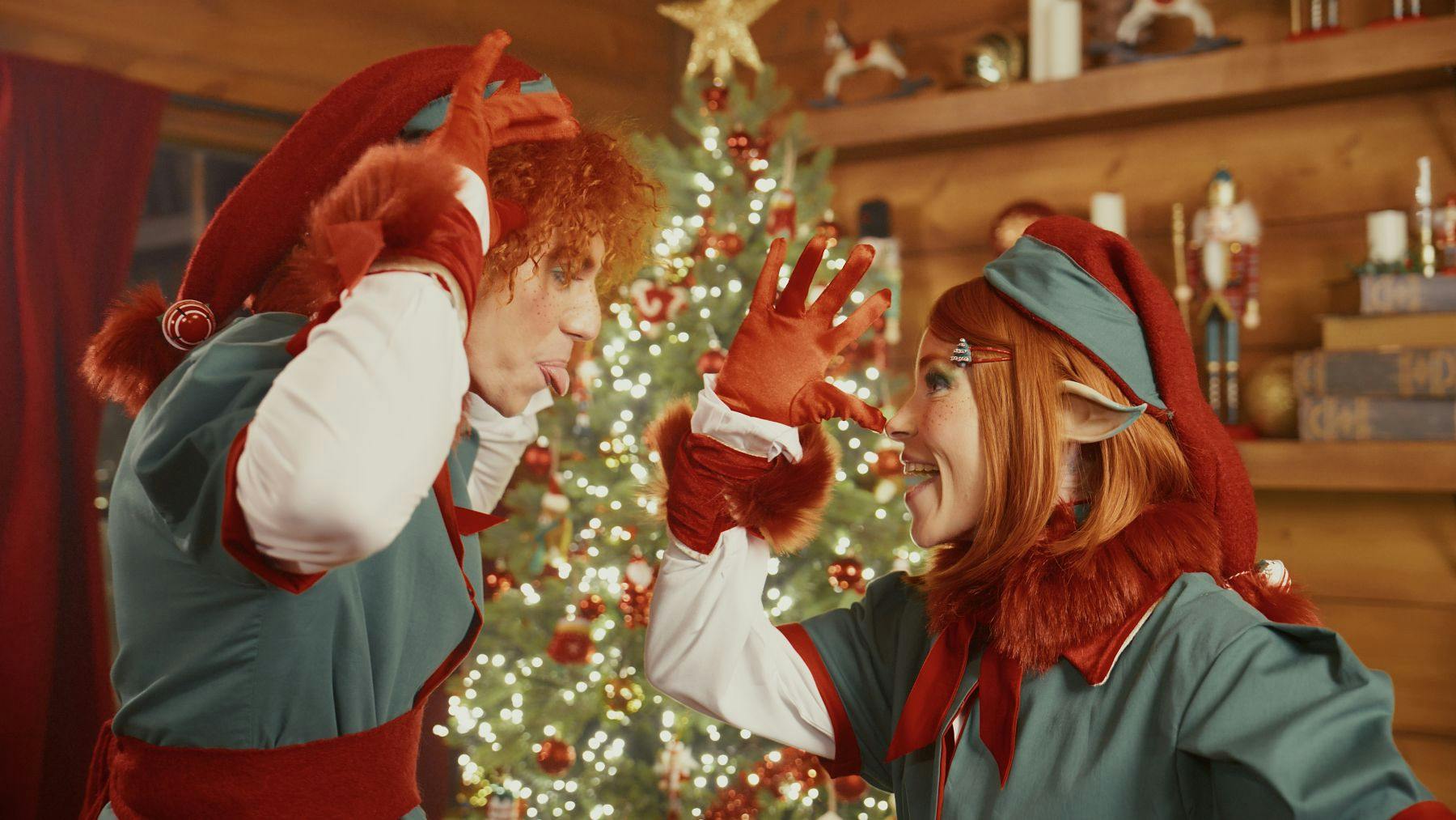 Two people dressed as Christmas elves, one touching the other's nose, with a decorated Christmas tree in the background.