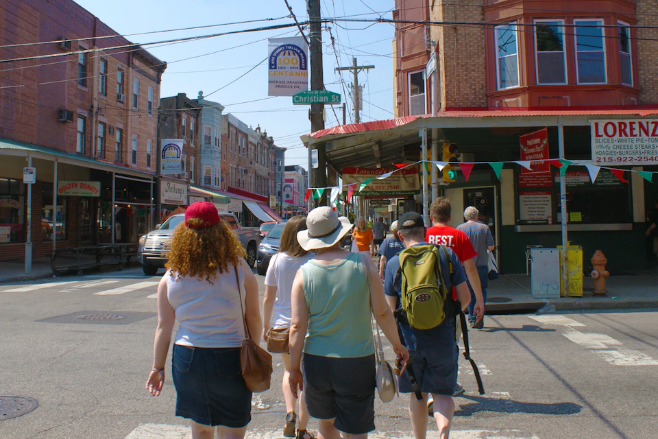 A group of people walking on a sunny street lined with brick buildings, colorful flags, and a street sign reading "Christian St."
