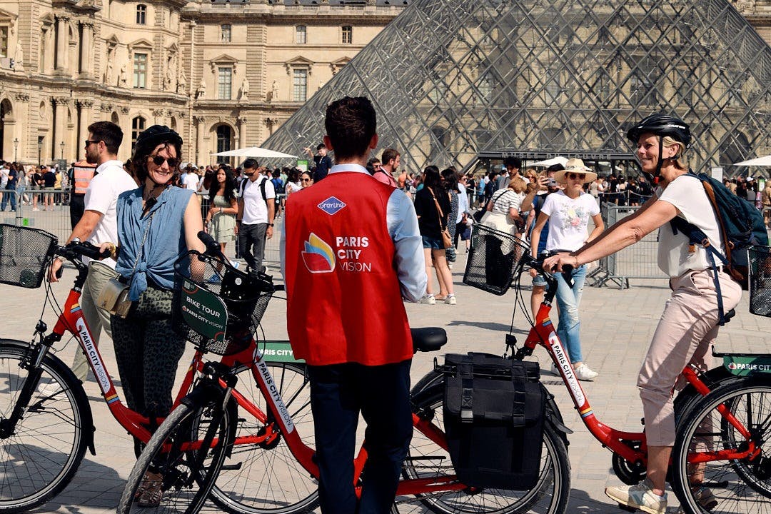 A tour guide in a red vest speaks to a cyclist near the Louvre Pyramid, surrounded by tourists.