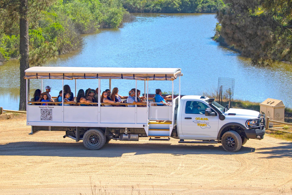 A group of people in an open-air tour truck driving on a sandy path alongside a body of water with greenery in the background.