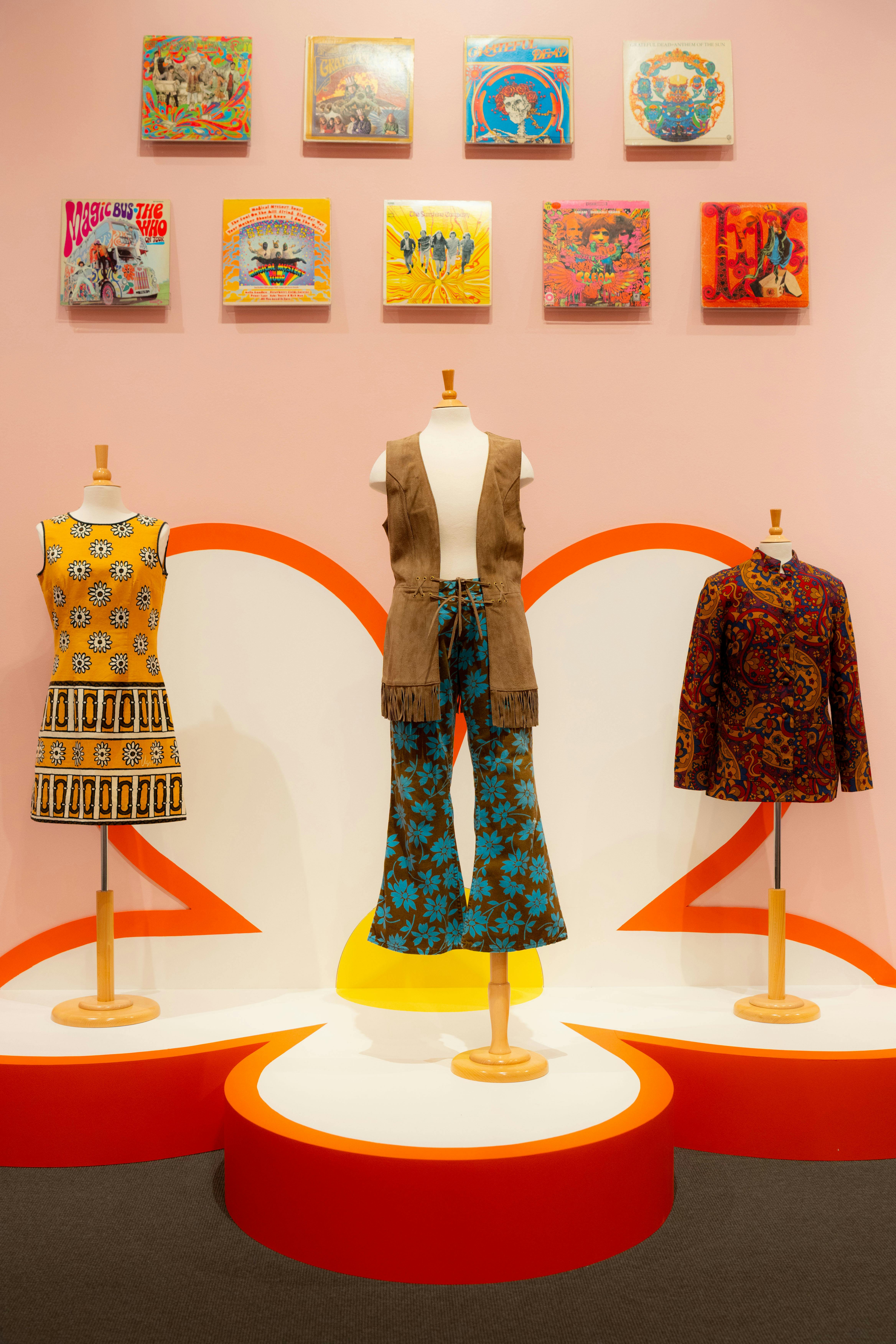 Tres maniquís vestits amb roba inspirada en la dècada de 1960 es mostren contra un colorit teló de fons amb pintures sobre ells.