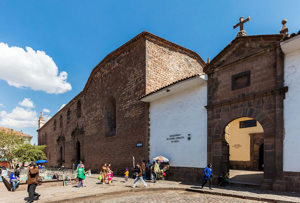 People walking near a historic stone building with arched windows and a small vendor stall under a bright blue sky.