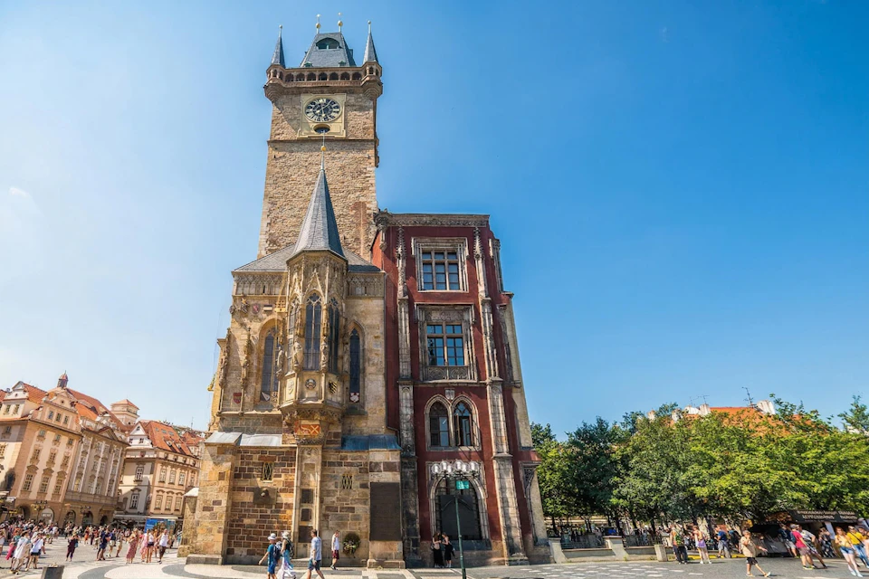 A historic clock tower and adjacent building under a clear blue sky, with people walking in the surrounding area.
