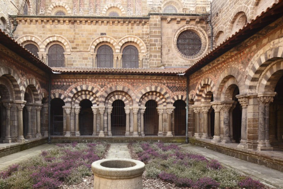 Medieval courtyard with stone arches and columns, featuring a central stone well and flowering plants in the foreground.