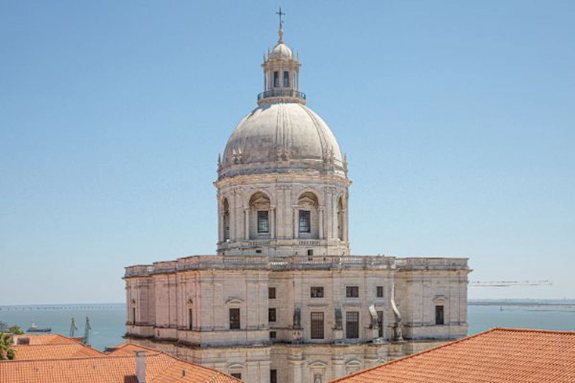 A large, white-domed building with arched windows and ornate details rises above terracotta rooftops against a clear blue sky.
