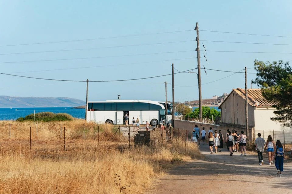 A group of people walk along a dusty path toward a large white bus parked near a coastal area with clear blue sky and power lines overhead.