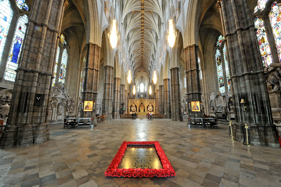 Interior view of a grand cathedral with high vaulted ceilings, chandeliers, and a memorial surrounded by red flowers on the floor.