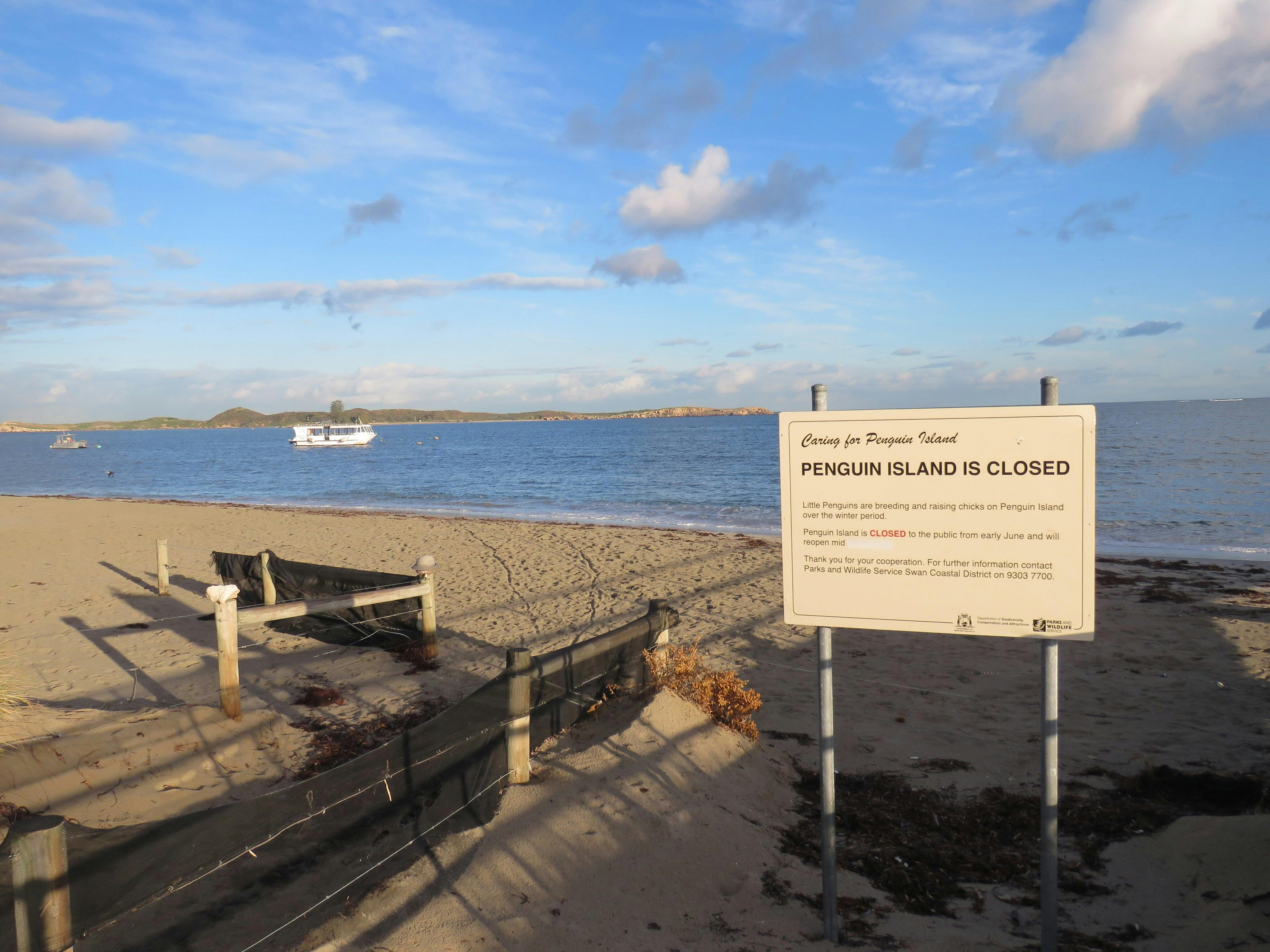 A beach with a sign stating "Penguin Island is closed." There is a boat on the water and a clear sky.