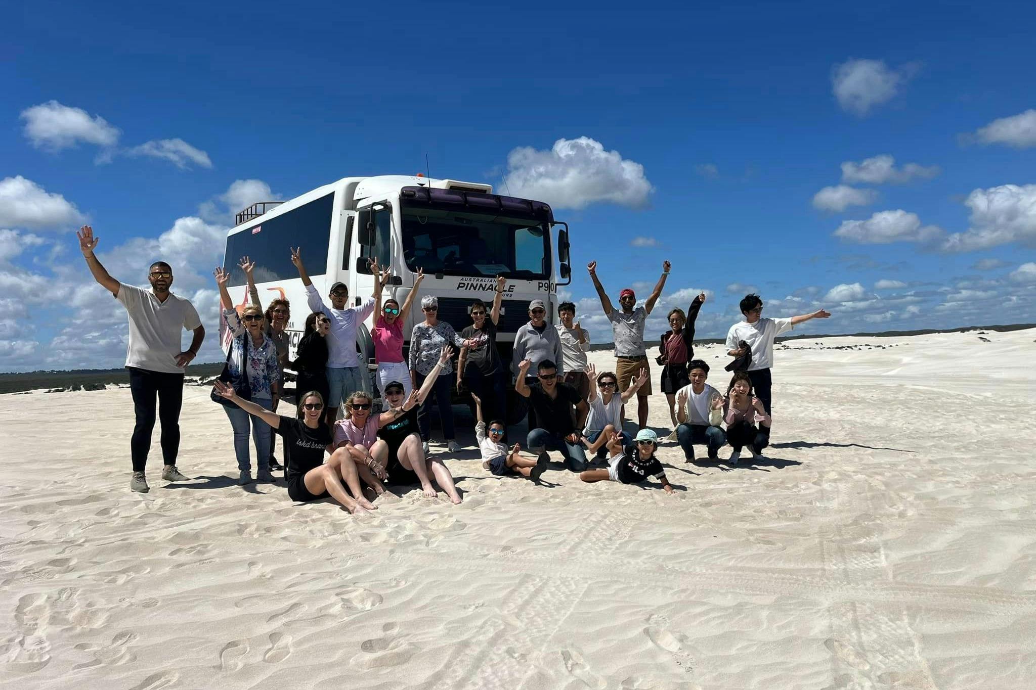 Een groep mensen die poseert voor een tourbus in een woestijnlandschap onder een blauwe lucht met hier en daar wat wolken.