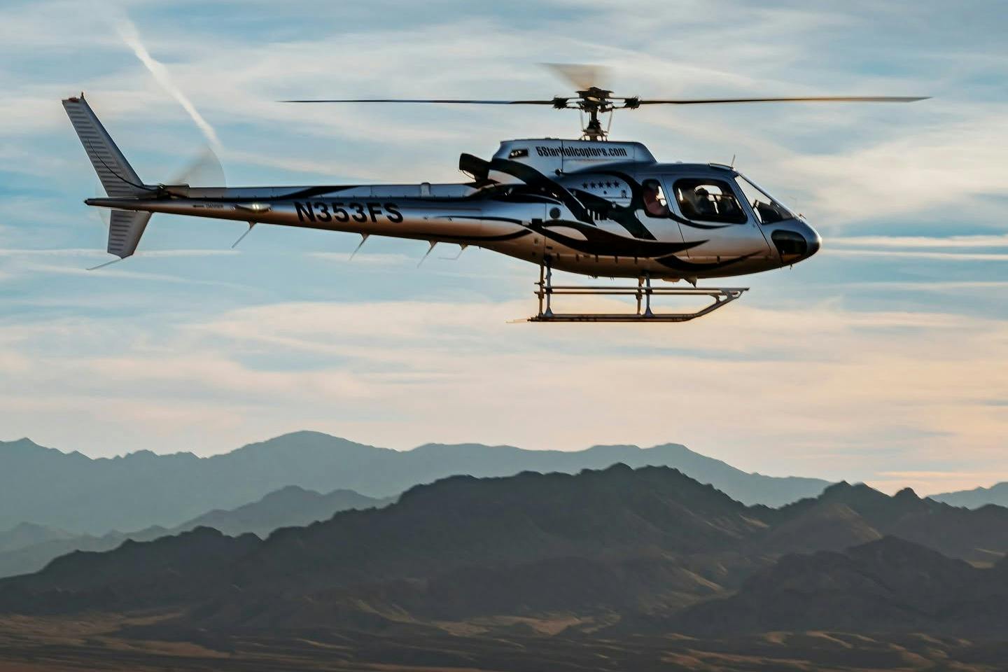 A helicopter in flight against a backdrop of mountains and a partly cloudy sky.