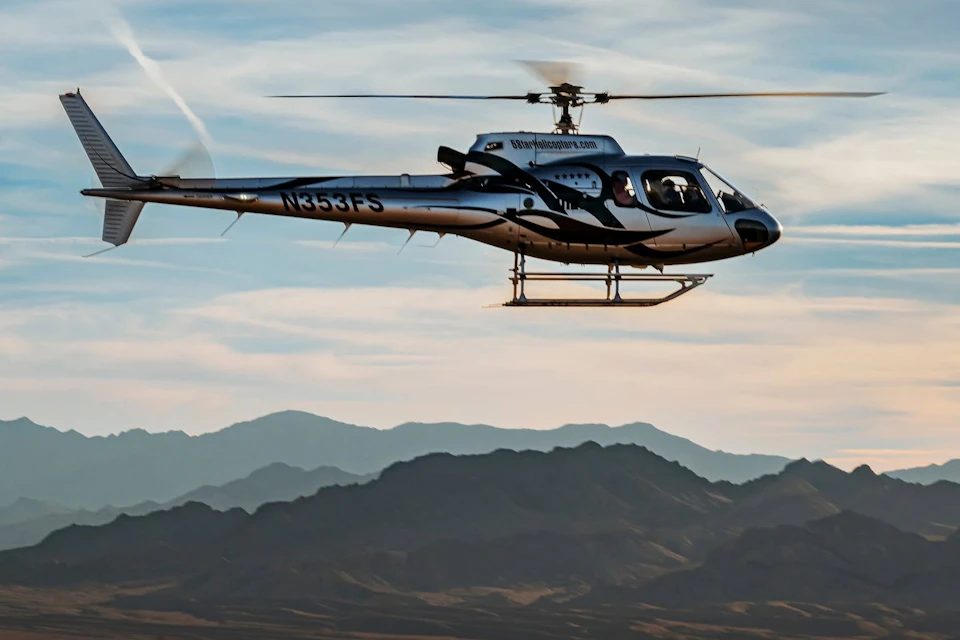A helicopter in flight against a backdrop of mountains and a partly cloudy sky.