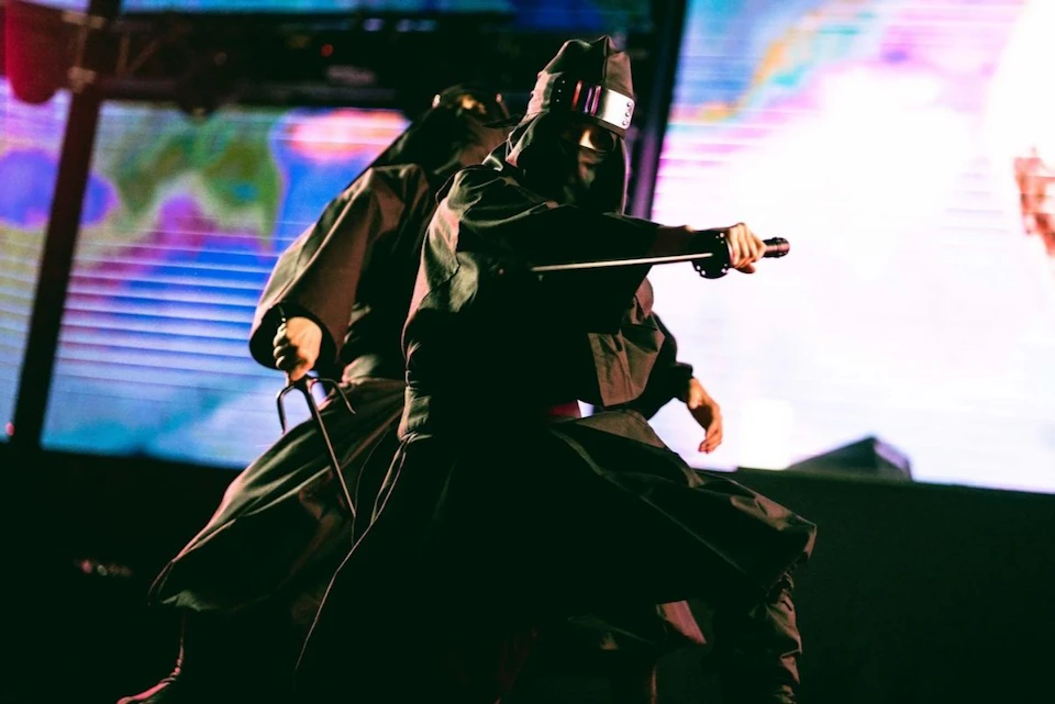 Two individuals in dark robes and hats perform a dramatic action pose with swords, against a colorful, abstract backdrop.