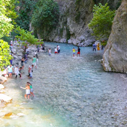 Människor vadar och njuter av en stenig, grund flod omgiven av träd och klippor en solig dag.