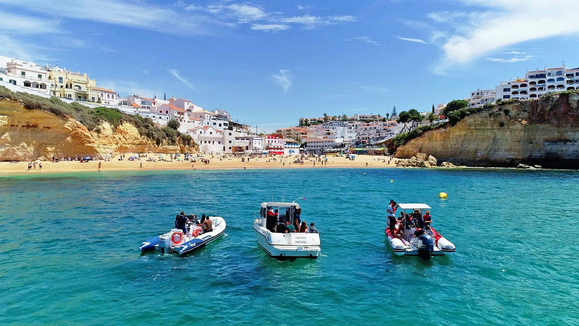 Three small boats with people are floating on a clear blue sea near a sandy beach with a town on a hillside in the background.