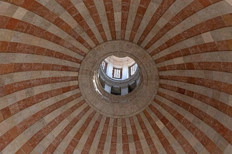 Looking up at a dome ceiling with alternating red and beige radial stripes and an oculus revealing a structure with windows and arches.
