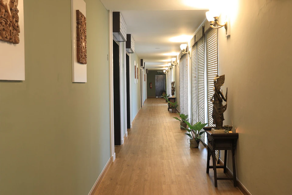 A hallway with wooden flooring, green walls, wall sconces, decorative statues, plants, and framed art pieces.