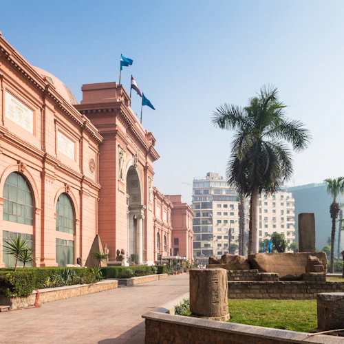 Ein großes rosafarbenes Gebäude mit Flaggen, architektonischen Elementen, Grünpflanzen, Palmen und antiken Artefakten im Vordergrund.