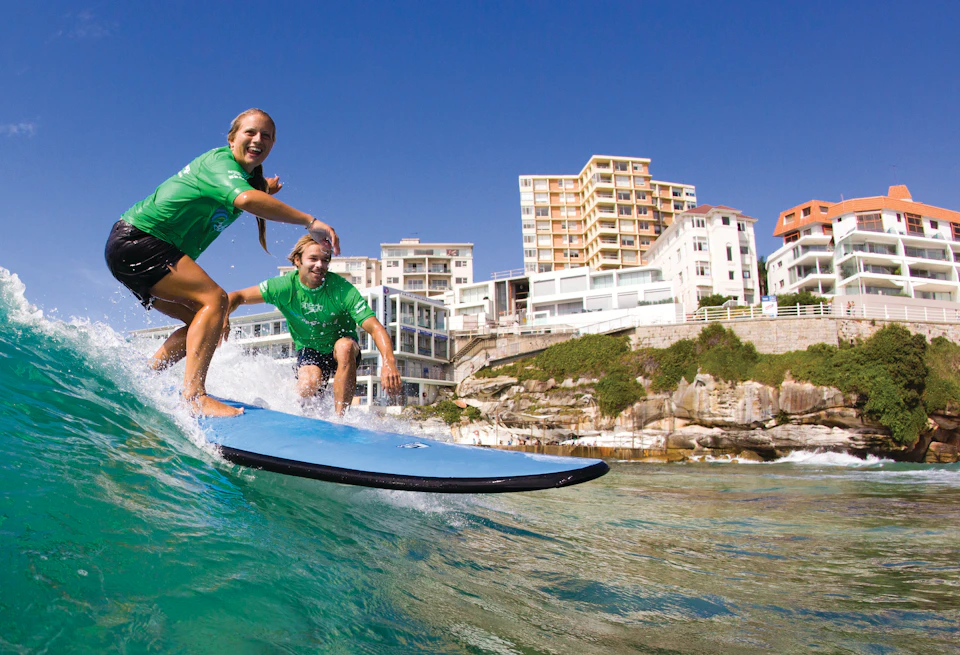Two people wearing green shirts ride a blue surfboard on a wave near a coastline with buildings in the background.