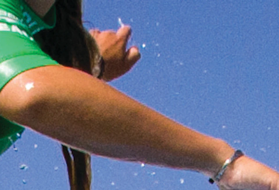 Two people wearing green shirts ride a blue surfboard on a wave near a coastline with buildings in the background.