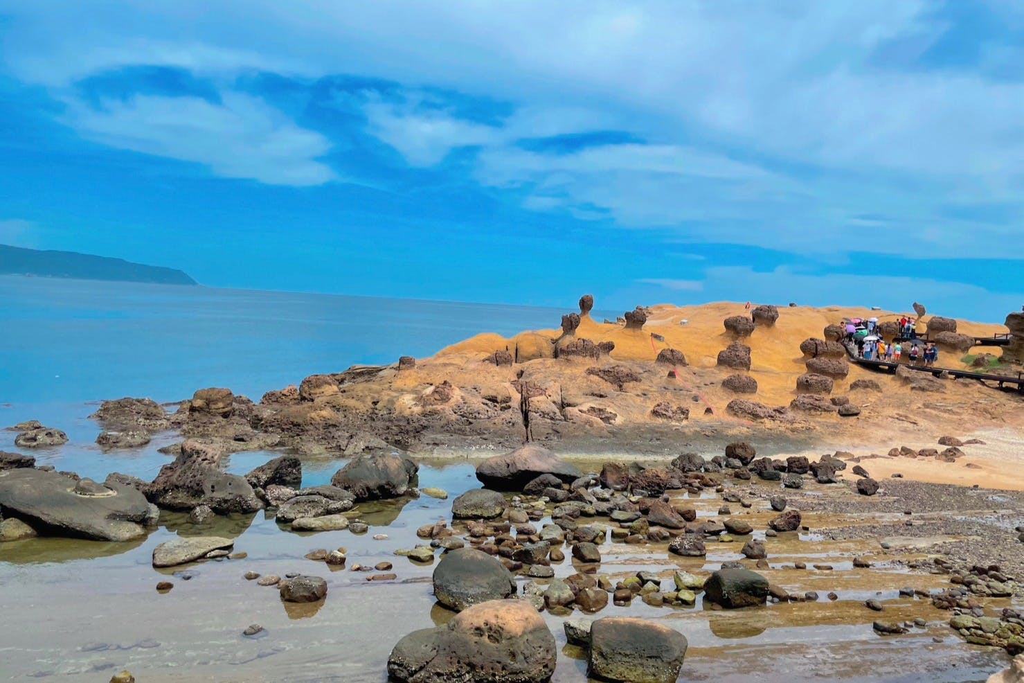 Rocky shore with unique rock formations extending into the sea under a bright blue sky with scattered clouds.