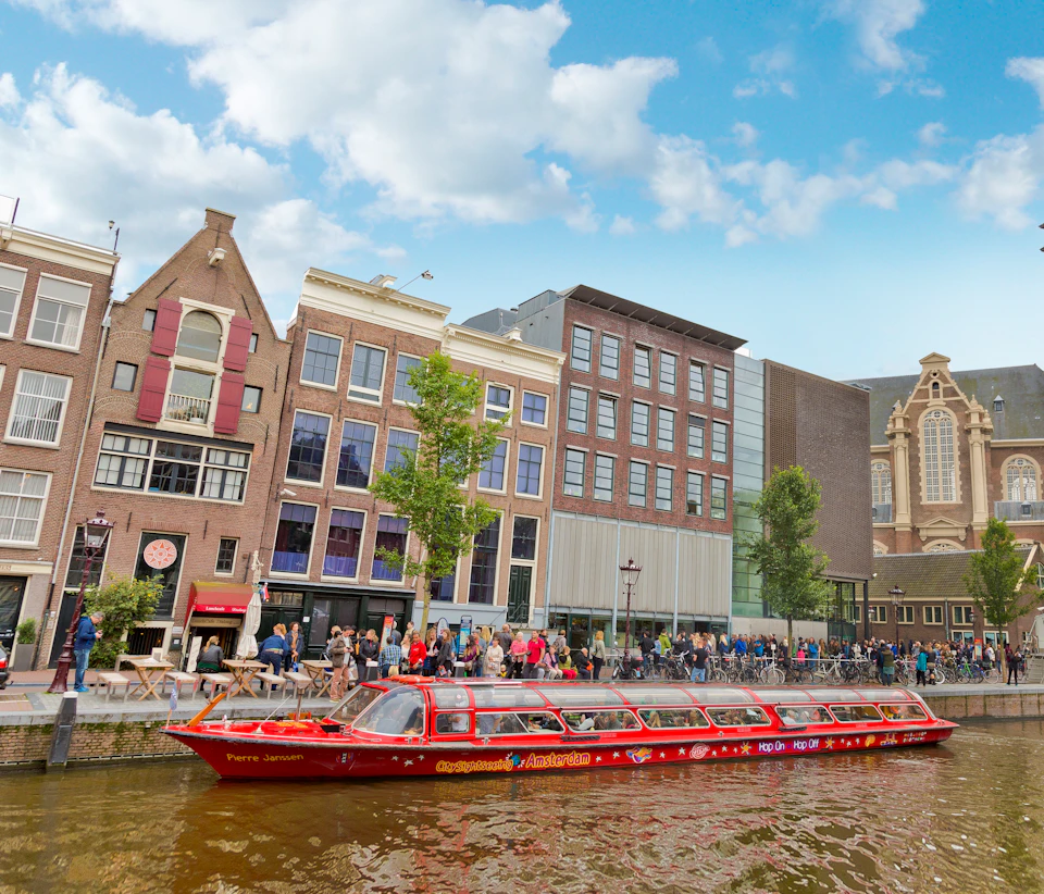 A red canal boat cruises by a row of traditional and modern buildings with people walking along the waterfront.
