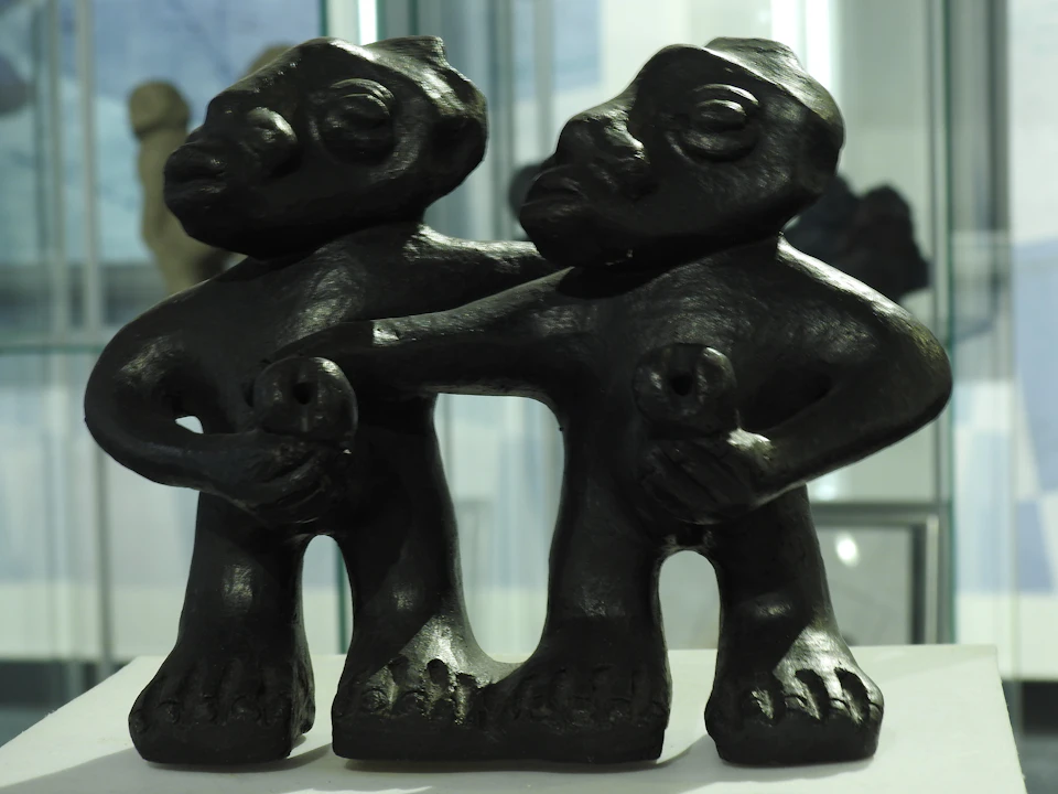 Two dark, twin-like abstract sculptures standing close together, each holding a small figure, set against a blurred background.