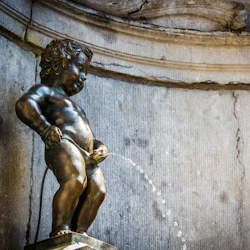 Bronsstaty av ett kissande barn, känt som Manneken Pis, mot en strukturerad stenmur.