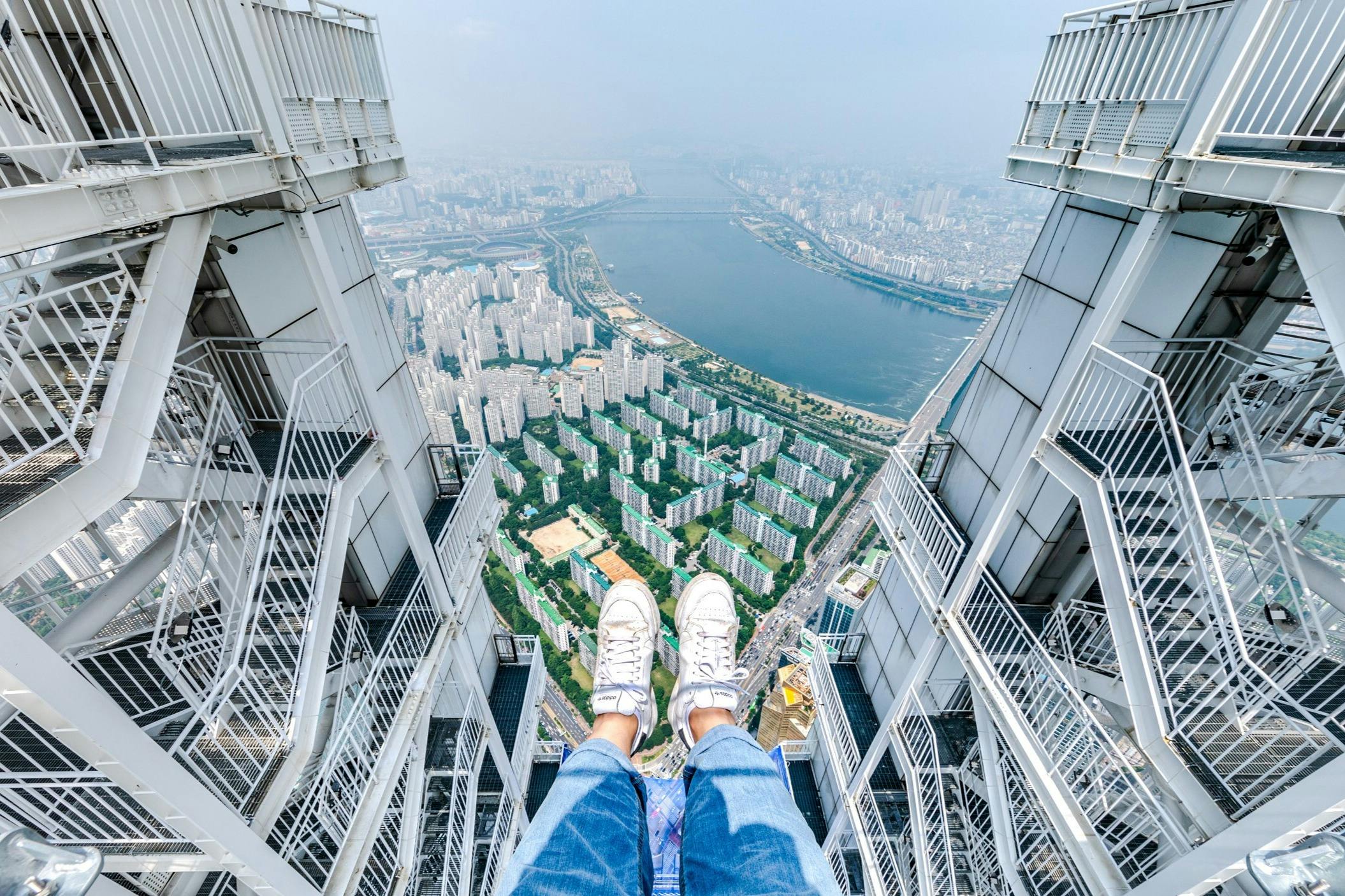 Pies en zapatillas y vaqueros colgando de un edificio alto, con vistas a un paisaje urbano con muchos edificios blancos y un río.
