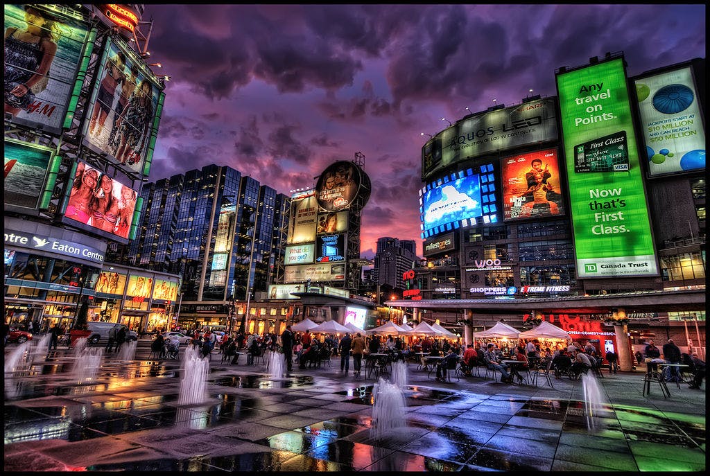 Crowded city square at sunset with bright electronic billboards, tall buildings, fountains, and tents.