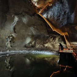 En person i en grotta står på en träplattform med utsikt över en reflekterande pool, med upplysta trappor uppför bergväggarna.