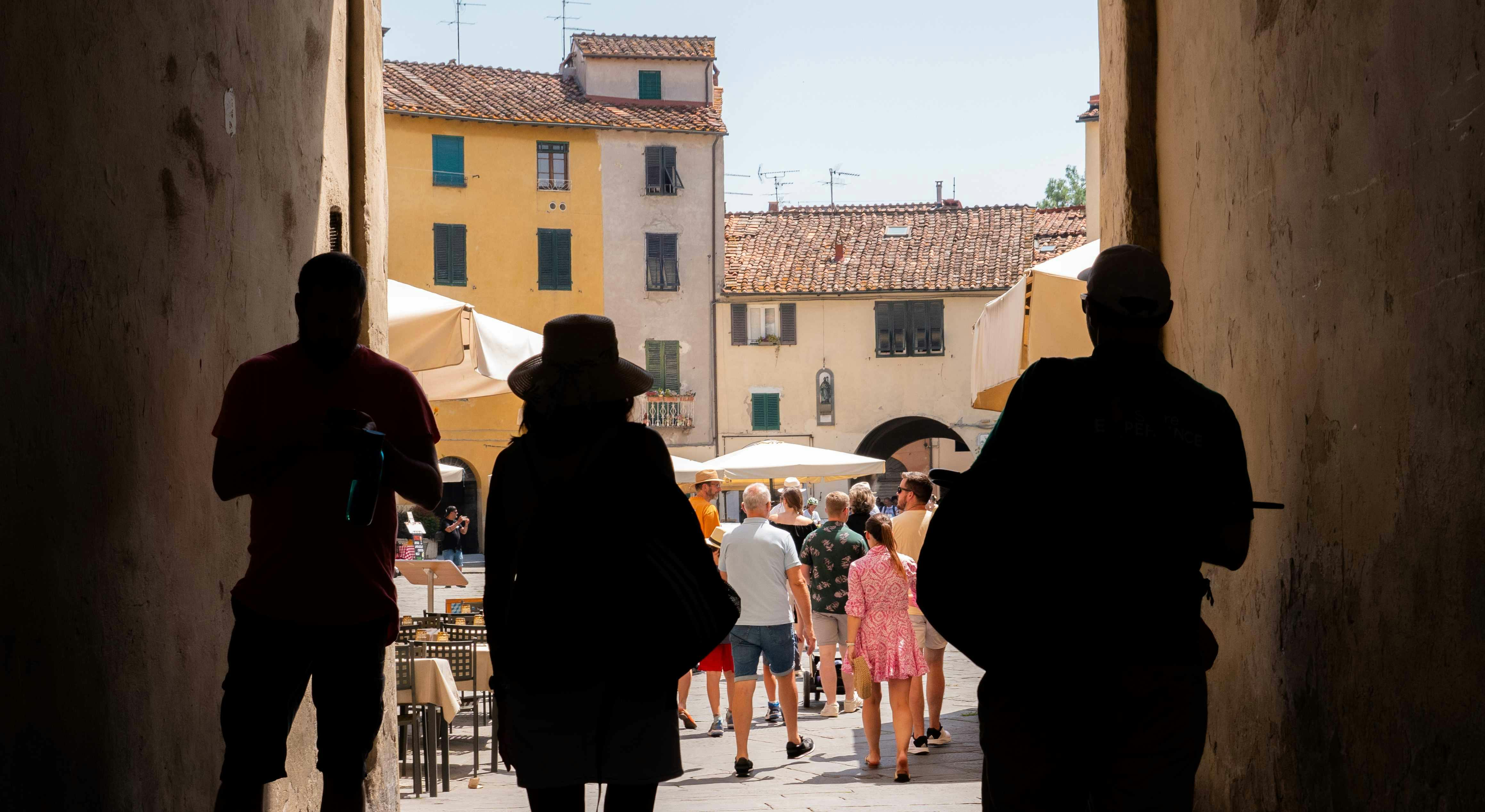 People walking through a Mediterranean street with old buildings, outdoor tables, and umbrellas, viewed from a shaded passage.