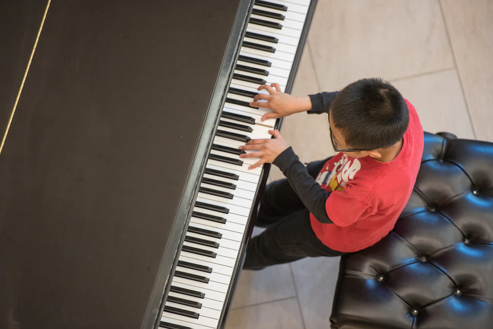 Child in a red shirt playing a large piano from a top-down perspective. Black and white keys are prominently visible.