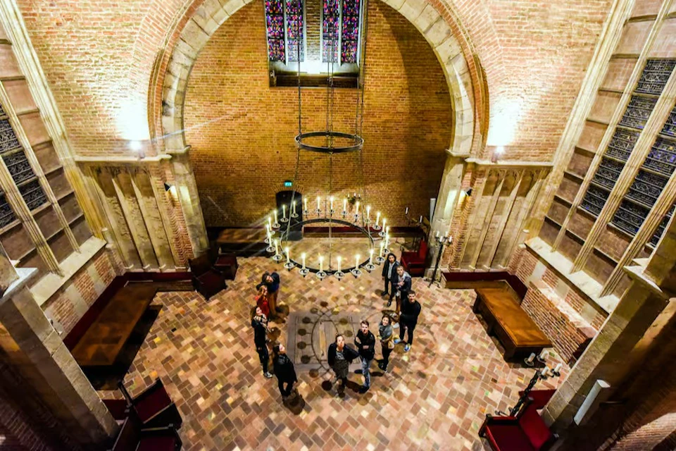A group of people standing in a spacious hall with brick walls, arched windows, and a large chandelier overhead.
