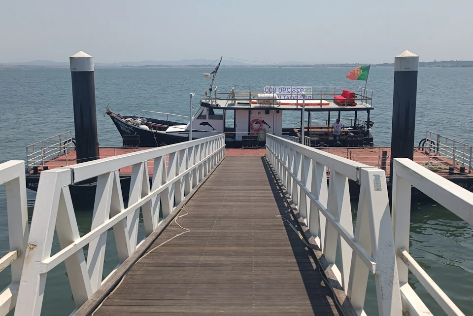 A docked tour boat at the end of a wooden pier, with a ramp leading to it. The boat has national flags and passengers on board.