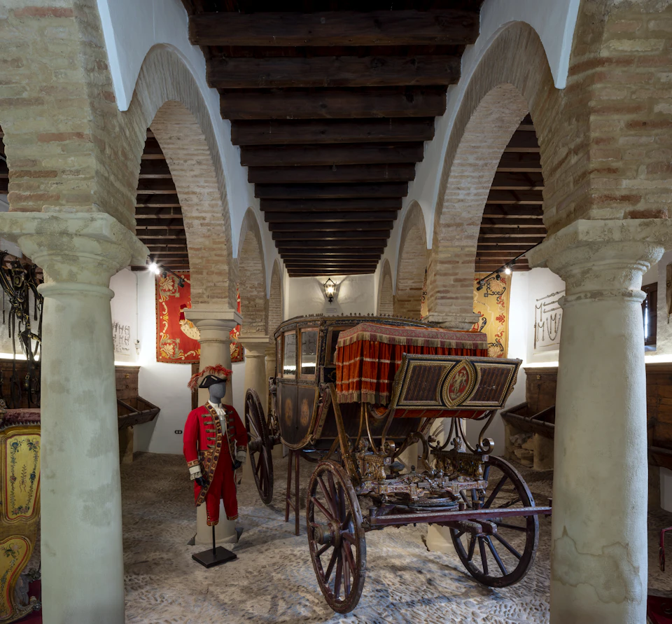 Historic carriage under an arched stone ceiling, flanked by tapestries, with a mannequin in period costume nearby.