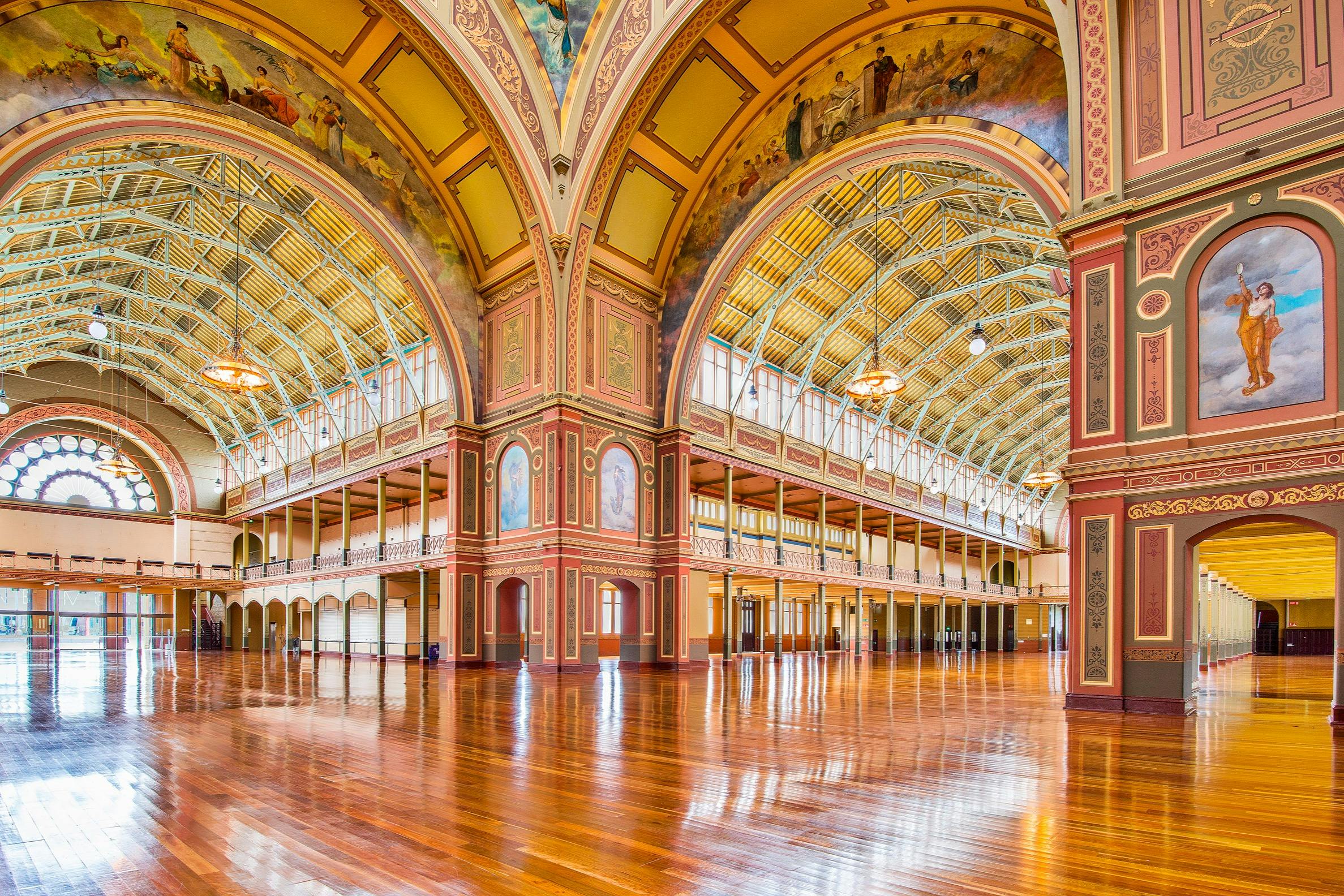 Gran salón ornamentado con techos arqueados, decoraciones intrincadas, suelos de madera y balcones, iluminado por lámparas de araña colgantes.