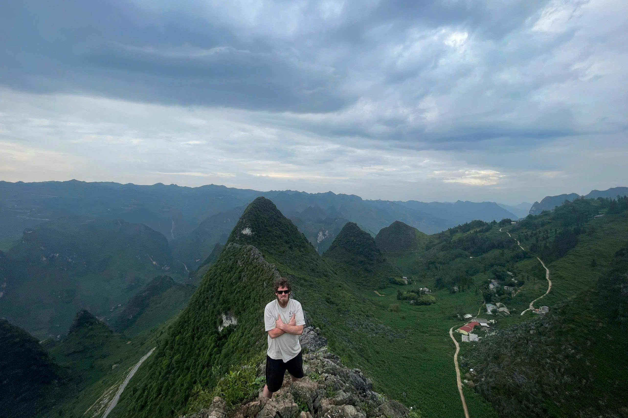 En person står på en klippetop med bjerge og overskyet himmel i baggrunden. Grønne dale og snoede stier er synlige nedenfor.