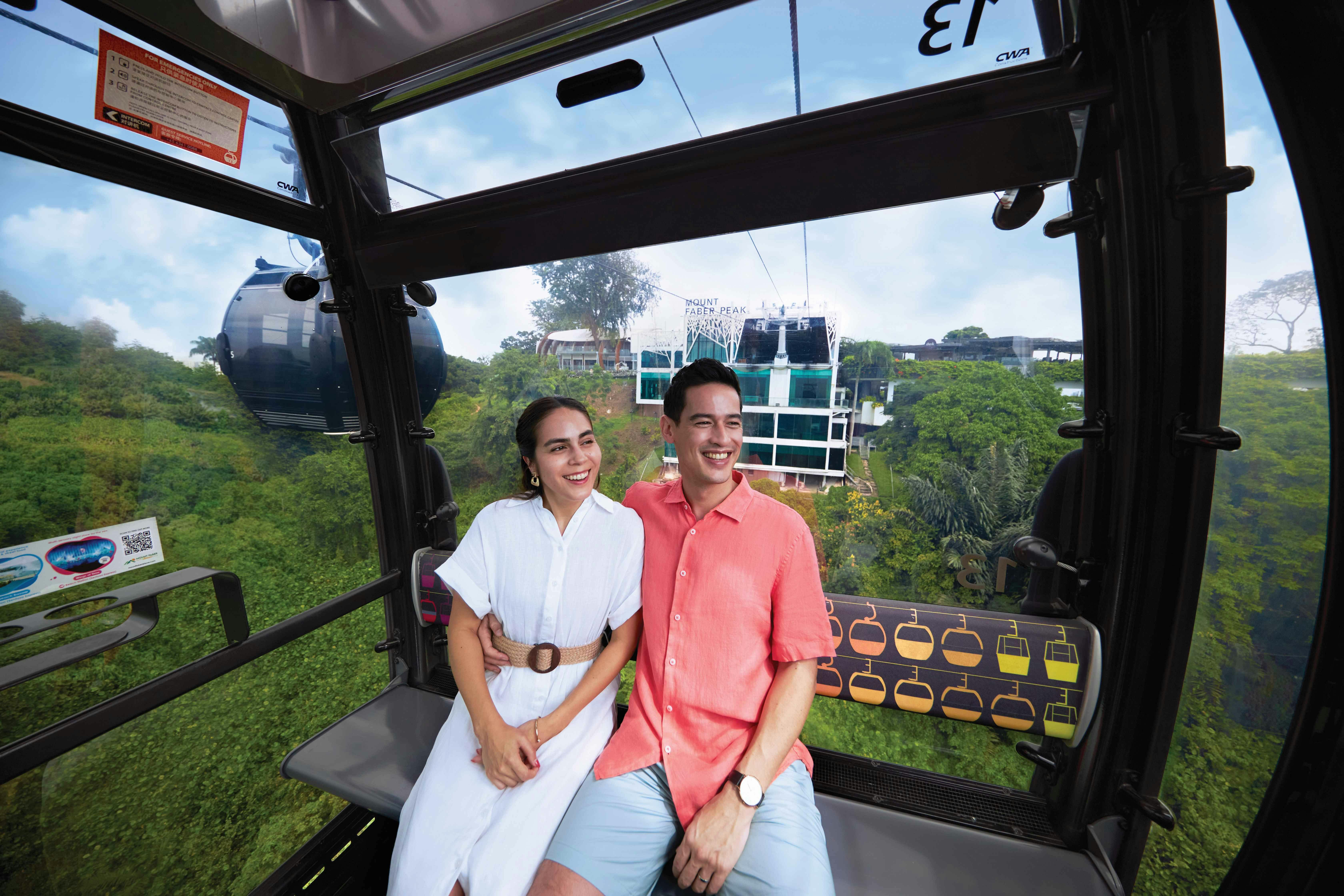 Ένα χαμογελαστό ζευγάρι κάθεται μέσα σε έναν ανελκυστήρα γόνδολας πάνω από ένα καταπράσινο τοπίο με κτίρια στο παρασκήνιο.