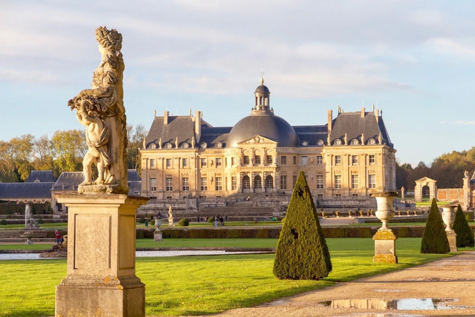A historic chateau with manicured gardens, statues, and a dome, lit by sunlight under a partly cloudy sky.