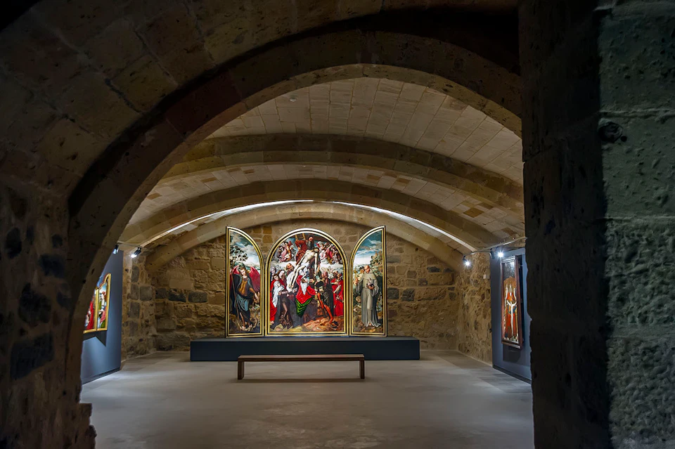 A stone-walled gallery room displaying a triptych painting and other artworks, with a wooden bench in the center.