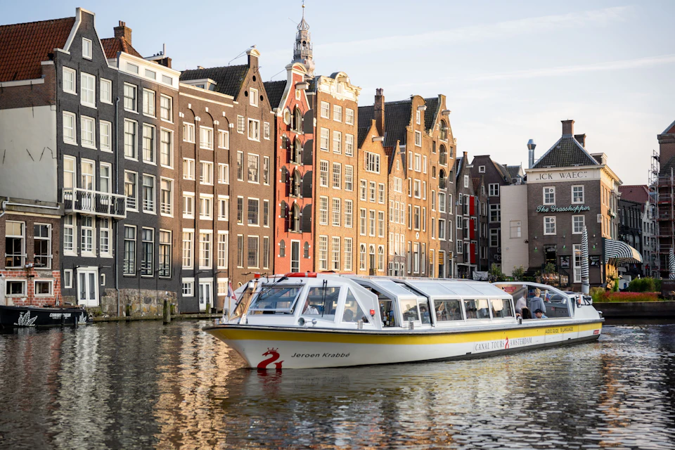 A canal boat passing by colorful, historic buildings along a waterfront in a city, under a partly cloudy sky.